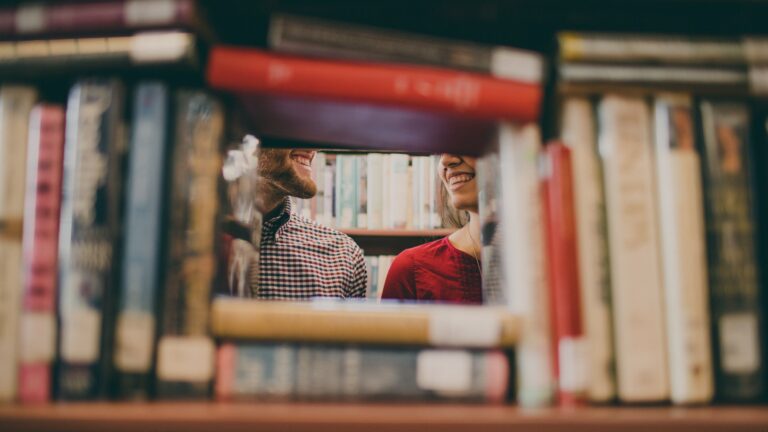 Two people smiling at each other through a gap in a bookshelf.